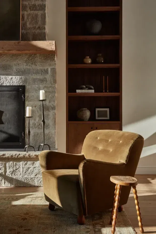 A warmly lit living room featuring a rustic stone fireplace with timber mantel, dark built-in wooden shelving and cabinetry, and a vintage caramel-brown tufted armchair beside a small turned-leg side table. Sunlight creates strong diagonal shadows across the neutral walls and timber floor, with a patterned rug grounding the seating area. The room has a cozy, traditional character with natural, earthy materials.