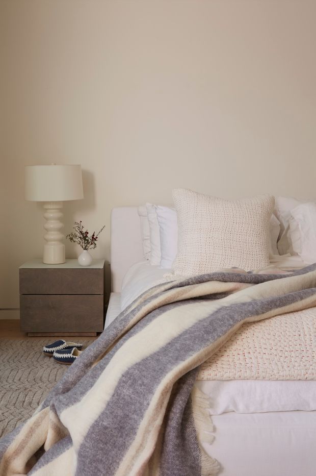 A softly styled bedroom with a low upholstered bed dressed in white bedding, a chunky cream knitted cushion, and a broad cream-and-grey striped throw. A dark wood bedside table holds a white ceramic lamp and a small vase of dried stems. The room has warm neutral painted walls, pale carpet, and a calm, layered textile palette.