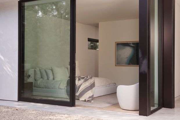 View into a calm, contemporary bedroom through large dark-framed glazed doors. The room contains a low upholstered bed with layered neutral bedding, a pale rounded lounge chair, a framed coastal artwork, light walls, and a soft neutral carpet. Reflections in the glazing show additional seating and greenery outside.