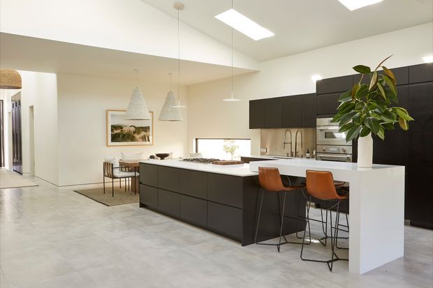 A bright, open-plan contemporary kitchen with a large white waterfall island and integrated breakfast bar, dark matte cabinetry, pale concrete-look flooring, and white walls. Two bar stools sit at the island, while a dining area with pendant lights is visible behind it. Built-in stainless-steel appliances, a sink, and a tall indoor plant complete the space.