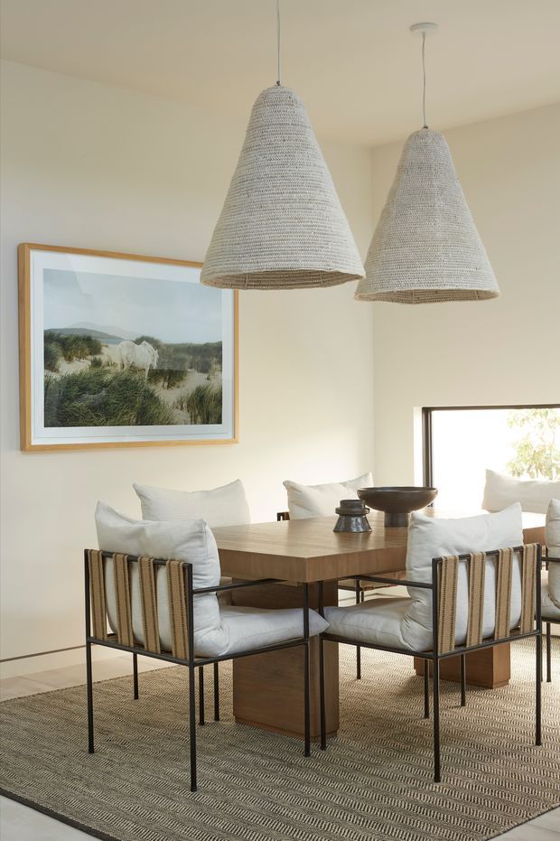 A softly lit dining room with a rectangular timber dining table surrounded by upholstered dining chairs with black metal frames. Two large woven pendant lights hang above the table, complemented by a textured neutral rug, cream walls, natural wood finishes, and coastal landscape artwork.
