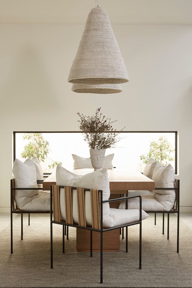 A refined dining room with a rectangular timber dining table surrounded by upholstered dining chairs with dark slim metal frames. A large woven pendant light hangs above the table, while a neutral textured rug, pale walls, and a broad dark-framed window create a warm, minimalist setting. A central vase with branches provides a subtle natural focal point.