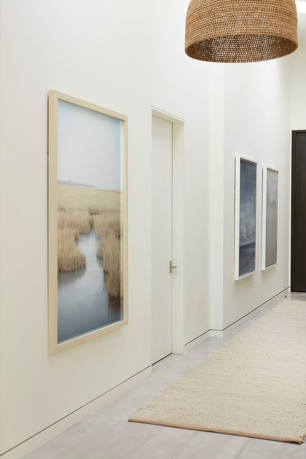 Bright minimalist interior hallway with white walls, flush white doors, pale flooring, framed landscape artwork, and a large woven pendant light. A neutral woven rug runs along the corridor, with additional framed artworks and a dark doorway at the far end.