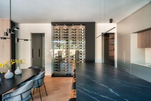 Contemporary open-plan kitchen and dining area with a large dark stone island benchtop, integrated pale wood cabinetry, timber flooring, and a prominent glass-fronted wine display cabinet. A dark dining table with upholstered chairs sits beside the kitchen, while black and neutral finishes create a refined, high-contrast interior.