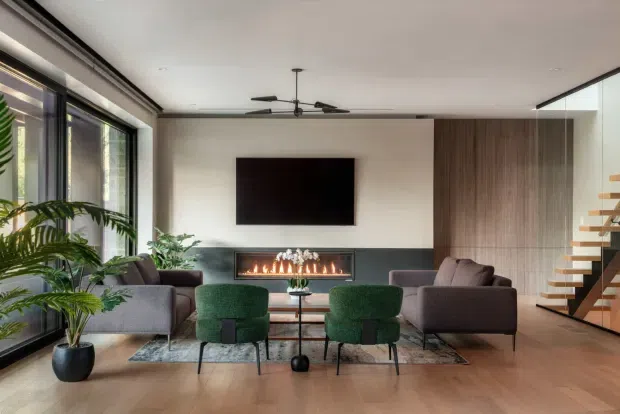 Contemporary living room with full-height glazing, a linear built-in fireplace beneath a wall-mounted television, green lounge chairs, grey sofas, timber flooring, and a floating timber staircase beside a vertical timber feature wall.