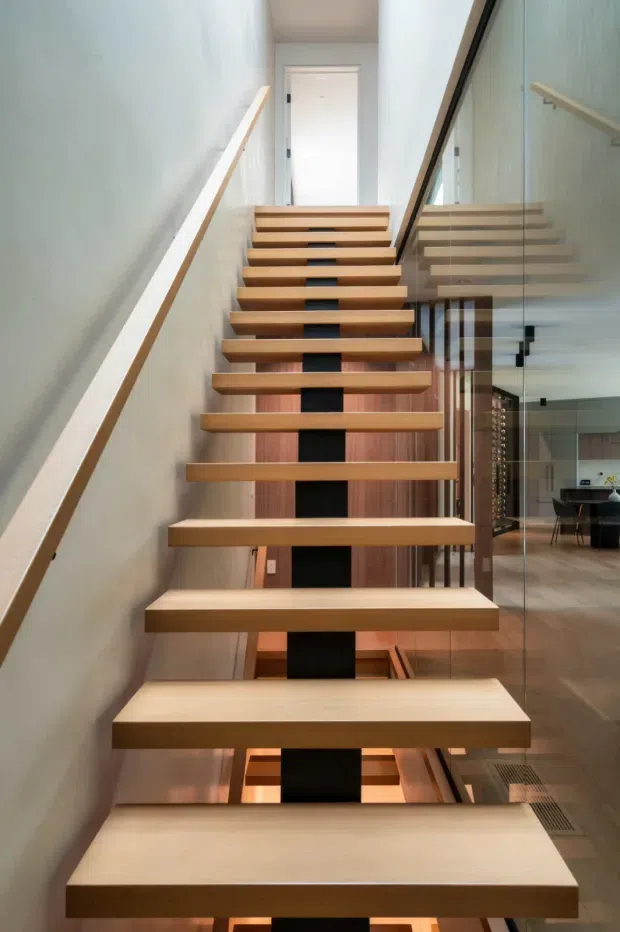 Interior view looking up a contemporary open-riser staircase with thick light timber treads and a dark central supporting spine. A clear glass balustrade runs along the right side, with a timber handrail on the left. The stairwell connects to an upper landing and overlooks a bright adjacent dining or living area visible through the glazing.