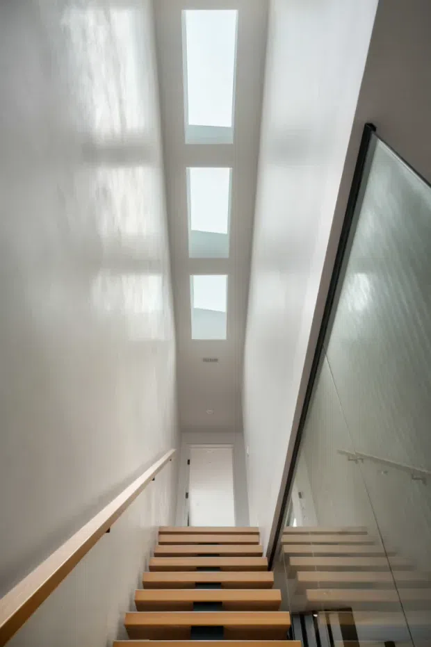 A narrow, bright contemporary stairwell viewed upward from the lower level, with floating timber treads, a simple timber handrail, white walls, a full-height glass balustrade, and three linear skylights illuminating the stair void. A white door is visible at the upper landing, and the glazing reflects the stairs and surrounding space.