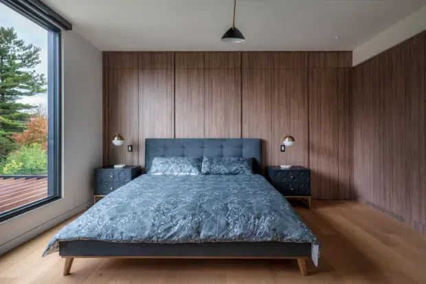 A refined bedroom with a low-profile upholstered bed centred against a full-height dark timber wall and matching timber floor. Integrated bedside tables, wall-mounted reading lamps, and a simple pendant light create a cohesive, minimalist composition. Large corner glazing brings in views of greenery and abundant natural light.