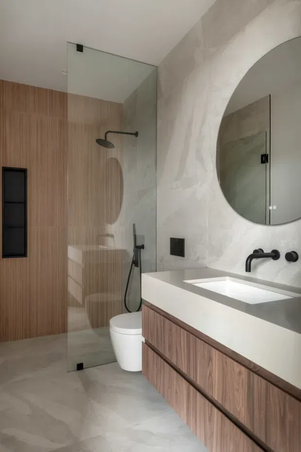 A refined contemporary bathroom featuring a walk-in shower enclosed by clear glass, a wall-hung toilet, a floating timber vanity with an integrated white basin, matte black wall-mounted tapware, a large round mirror, and warm vertical timber wall panelling. The remaining walls and floor have a pale stone-like finish, creating a calm, warm-toned interior.