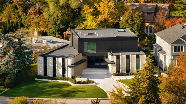 A contemporary multi-level residence viewed from above, set within a leafy suburban neighbourhood. The home features a dark box-like upper volume, pale masonry walls, extensive glazing, flat and low-pitched roof forms, a landscaped front lawn, and a broad paved entry approach. Mature autumn trees and neighbouring houses surround the property.