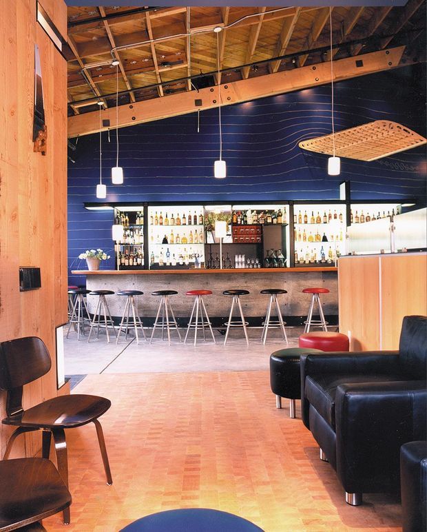 Interior view of a lively bar or hospitality venue with a long backlit bottle display and timber-fronted bar, arranged with a row of metal bar stools. The space features a dramatic dark blue textured feature wall, exposed timber roof structure and services, suspended pendant lights, warm timber parquet flooring, and black leather lounge seating in the foreground. The overall aesthetic is eclectic and industrial with strong mid-century influences.