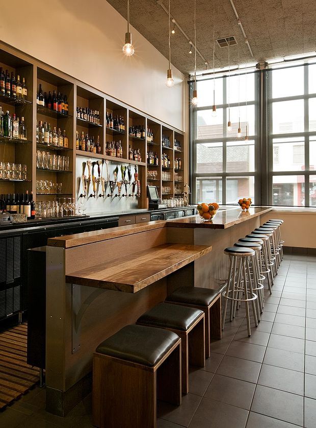 A spacious commercial bar interior with a long timber counter and integrated seating, extensive back-bar shelving displaying bottles and glassware, beer taps, refrigeration, and a second bar counter lined with stools. Large industrial-style steel-framed windows provide abundant daylight. The ceiling has an exposed textured finish with suspended track lighting and pendant bulbs, while the floor is finished in large grey tiles.