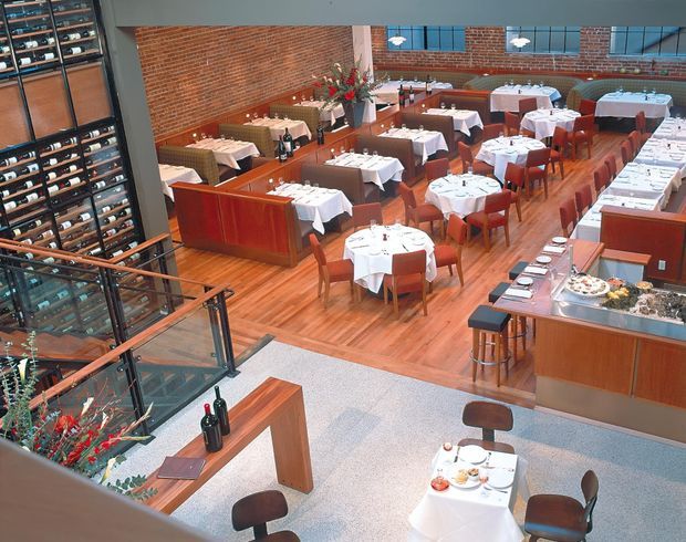Elevated view of a spacious restaurant dining room with numerous white-clothed tables, red upholstered chairs, warm timber flooring, exposed red-brick walls, and substantial timber service counters. A tall wine display and cellar wall occupies the left side, while a glass-and-metal stair or balcony railing overlooks the dining area. The space has a refined, warm hospitality interior with abundant natural light.