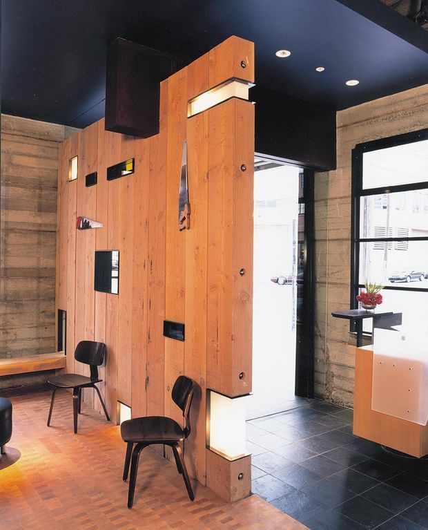 Interior view of a dark, wood-lined commercial or institutional space featuring a sculptural vertical timber partition and built-in display niches. The partition incorporates integrated lighting and storage-like openings, with black modern chairs, a bench, black-framed windows, and a tiled entry zone. Exposed rough concrete-block walls and a black ceiling create an industrial atmosphere.