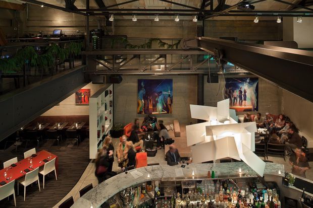 A dramatic multi-level industrial-style hospitality interior, viewed from an elevated position over a busy bar and dining area. Exposed steel beams, concrete walls, dark metal walkways and railings, track lighting, large colourful artworks, indoor greenery, and a sculptural cluster of white pendant lights define the space. A curved bar with extensive bottle displays occupies the foreground, with dining tables, booths, and patrons distributed across the lower level and mezzanine.