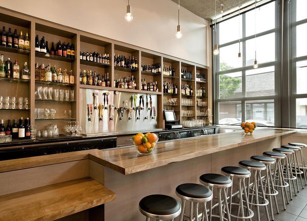 A bright, contemporary commercial bar interior with a long timber-fronted counter and extensive built-in shelving stocked with bottles and glassware. Beer taps, bar equipment, pendant bulbs, and a row of black-cushioned metal stools define the space. Large industrial-style grid windows bring in abundant daylight, while bowls of oranges add small decorative accents.