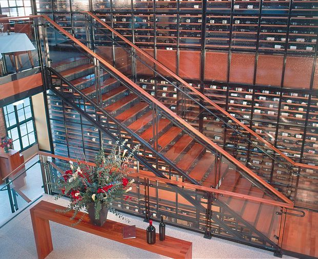 A multi-level interior atrium dominated by a broad staircase with dark metal structure, timber handrails, and transparent glass balustrades. Behind the stairs is an extensive wall of dark timber and glass shelving or display storage, giving the space a library-like or commercial character. Warm reddish-brown timber accents contrast with pale flooring and large glazed windows. A built-in timber console with decorative objects and floral arrangements sits in the foreground.