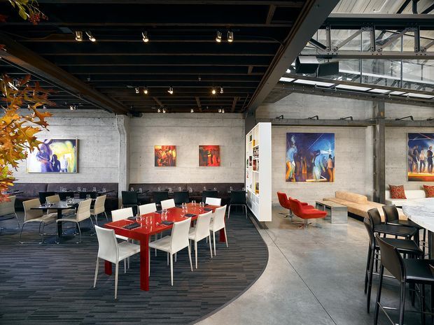 Open-plan contemporary commercial interior with a dining and lounge arrangement. The space features exposed concrete walls and columns, a dark exposed structural ceiling with track lighting, polished concrete flooring, dark striped carpet, modern red and white dining tables and chairs, black banquet seating, a lounge area, and colorful artwork. Large overhead glazing and visible steel framing bring natural light into the industrial-style space.