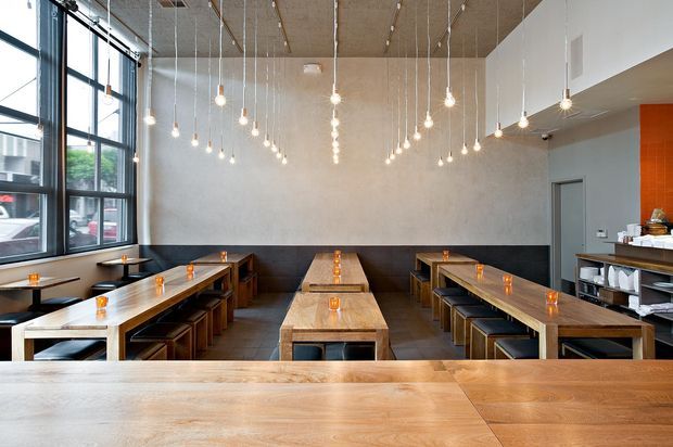 A modern commercial dining space, likely a café or casual restaurant, with long communal timber tables, integrated bench seating, dark upholstered banquettes, and a service counter at the right. Floor-to-ceiling industrial-style black-framed windows line the left wall, while numerous pendant lights hang from a timber ceiling. The restrained palette of timber, charcoal, grey, and warm orange accents creates a contemporary, welcoming atmosphere.
