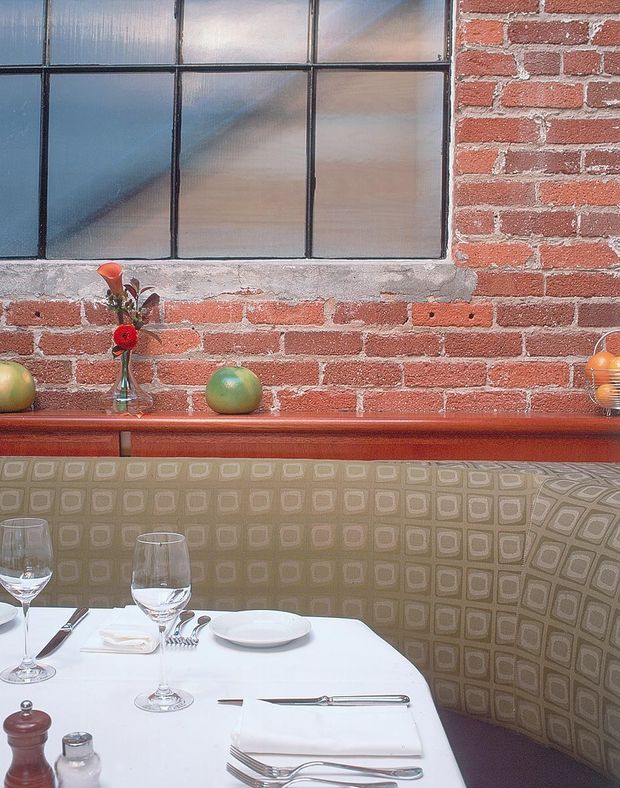 Close-up interior view of a restaurant-style dining booth. A patterned upholstered banquette wraps around a white-clothed dining table set with plates, wine glasses, cutlery and folded napkins. Behind it is an exposed red brick wall with a large industrial-style multi-pane frosted window, a timber ledge, and small decorative fruit and flowers. The overall feel is warm, intimate and vintage-industrial.