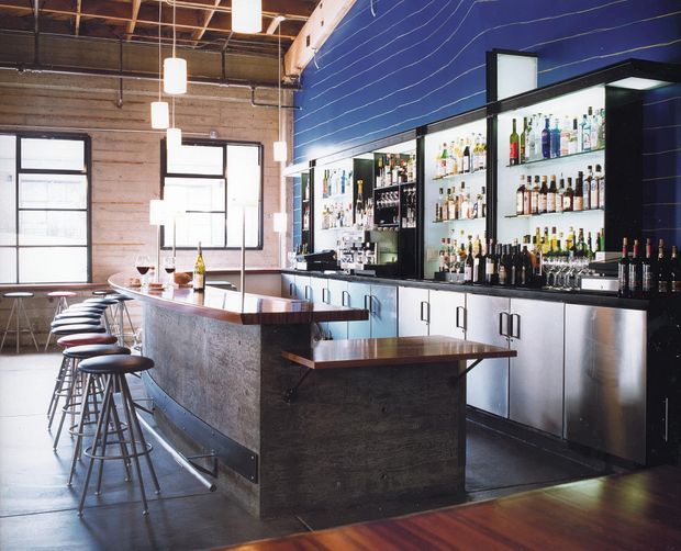 Interior photograph of a modern industrial-style bar or hospitality venue. A long concrete-fronted bar counter with dark timber tops and rows of black metal stools faces a back bar with extensive bottle shelving, stainless-steel cabinetry, and integrated refrigeration. The space has exposed timber roof framing, concrete walls, large black-framed windows, pendant lights, polished dark flooring, and a deep blue feature wall with horizontal line detailing.