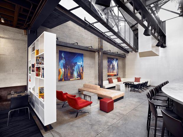A large open-plan industrial-style interior with polished concrete floors and raw concrete walls, exposed dark steel beams, visible services and a glazed roof structure. The space combines a lounge area with red lounge chairs, a low upholstered bench and ottoman, artwork, a freestanding white shelving partition, and a dining or meeting area with a long table and black chairs. High-level glazing and skylights create a bright, dramatic atmosphere.