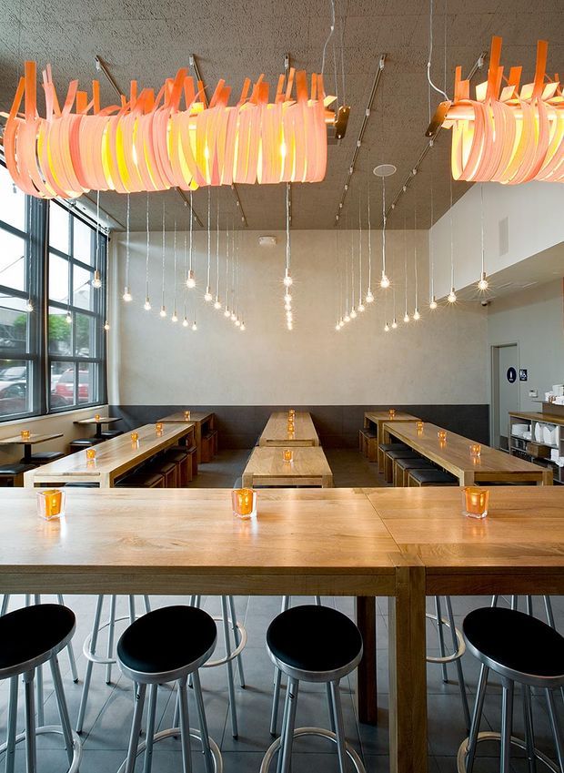 Interior view of a contemporary casual dining venue with long communal timber tables, black bar stools, exposed concrete ceiling, large industrial-style windows, and numerous hanging pendant lights. A large sculptural orange and pink linear light fixture dominates the ceiling, while small warm table lights create an inviting atmosphere.