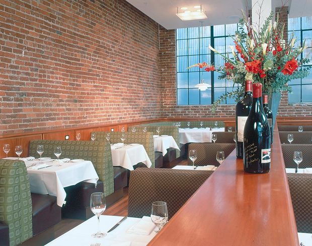 Interior view of an upscale restaurant dining room with exposed red-brick walls, large black-framed industrial-style windows, patterned upholstered banquette seating, neatly dressed white tablecloth tables, and a prominent polished wood service or bar counter in the foreground. Wine bottles and a floral arrangement form a centerpiece. The space has a warm, refined industrial character with abundant natural light.