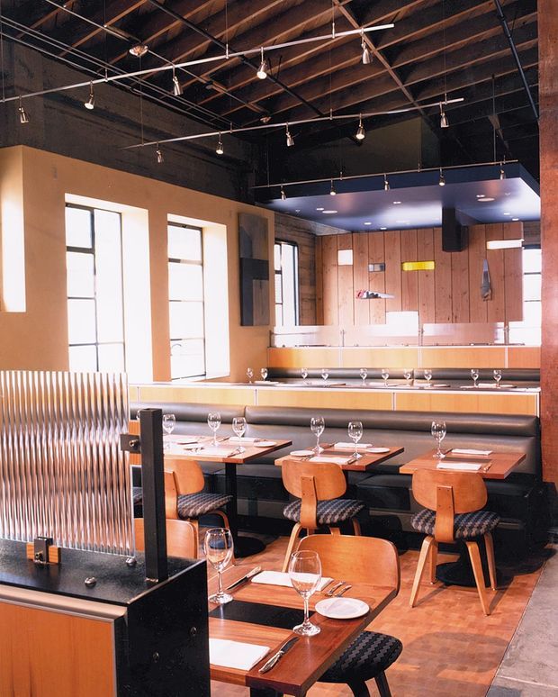 A warmly lit restaurant dining room with rows of timber tables, upholstered banquette seating, and wooden dining chairs. The space features a high exposed timber and metal ceiling with suspended track and pendant lighting, tall industrial-style windows, dark painted walls, timber wall panelling, and a long service or bar counter at the rear. A ribbed metal privacy screen and dark stone or laminate host counter occupy the foreground.