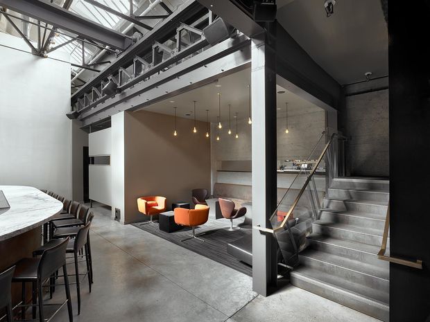 A dramatic industrial-style interior featuring an open-plan dining and living area beneath exposed structural steel and a glazed roof. A long dining table with dark chairs occupies the left foreground, while a sunken lounge with brightly coloured chairs and pendant lights sits centrally. Concrete flooring, dark steel beams, exposed services, and a concrete stairway with metal balustrade reinforce the warehouse-like character.