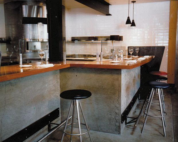 A compact industrial-style kitchen and breakfast bar with a concrete island base, warm timber benchtop, stainless-steel appliances, glossy white tiled splashback, black structural elements, and pendant lighting. Several black bar stools line the raised counter, which is set for dining with glassware and place settings.
