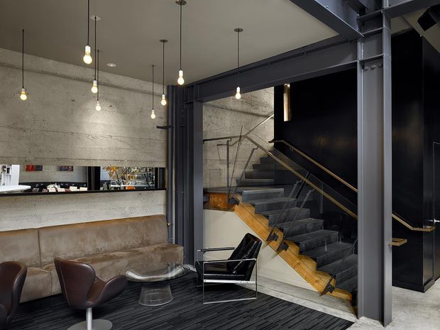Industrial-style interior lounge and stair hall with exposed dark steel beams and columns, raw concrete walls, blackened metal staircase, timber stair treads and handrail accents. A leather sofa, lounge chairs, glass coffee table, and suspended bare-bulb pendant lights occupy the foreground, while a bar or service counter is visible through the opening at left.