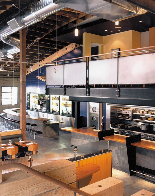 A spacious multi-level commercial restaurant and bar interior with an open industrial fit-out. The image shows a visible commercial kitchen with stainless-steel equipment, a bar with bottle displays and stools, dining tables, booth seating, a mezzanine or raised platform with metal railings, exposed timber beams, black ceilings, and prominent exposed spiral HVAC ductwork. Warm timber surfaces contrast with dark metal, concrete-like finishes, and translucent partition panels.
