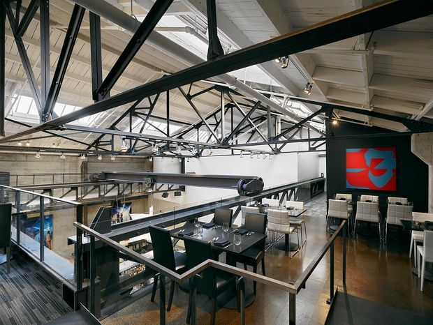 An elevated view of an industrial-style commercial interior, likely a restaurant or hospitality venue. The space features exposed steel roof trusses, white-painted structural beams and ceiling panels, concrete and dark painted walls, polished dark flooring, black dining tables and chairs, and extensive internal glazing and metal balustrades. Track lighting and a large red-and-blue graphic artwork add visual accents, while the open mezzanine-like arrangement overlooks the lower level.