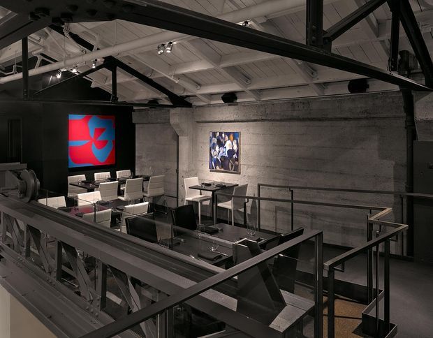 An industrial-style commercial interior viewed from an elevated mezzanine or walkway. The space features exposed black steel trusses and beams, a white painted industrial ceiling with track lighting and visible services, rough grey concrete walls, metal balustrades, and a lower-level meeting or dining area furnished with tables and chairs. Artwork and a bright digital display add colour to the otherwise monochrome setting.