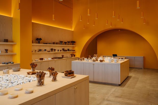 A warm monochromatic ochre-orange retail or display interior with pale timber cabinetry, long open display shelves, central island counters, ceramic objects and sculptural vessels. The space features a large rounded arched wall opening, polished concrete or stone-look flooring, and numerous small suspended pendant lights.