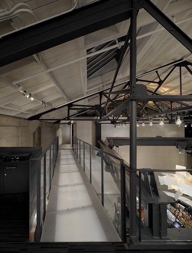 Interior view of a multi-level industrial-style commercial space, featuring an elevated metal walkway with glass and black metal balustrades, exposed black steel trusses, a white painted structural ceiling, concrete walls, track lighting, and a bar or display area below.