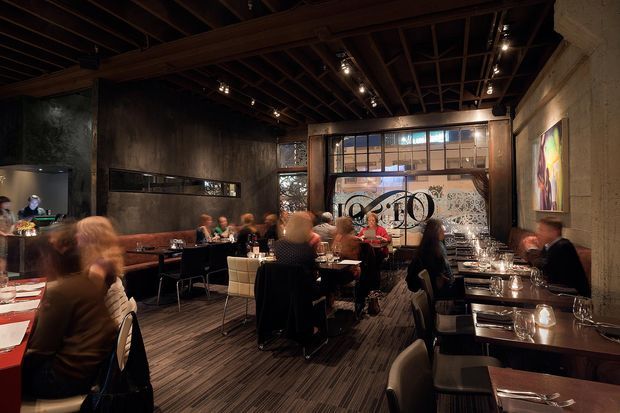 A dimly lit, industrial-style restaurant dining room with exposed dark timber ceiling beams, concrete-textured walls, dark timber flooring, black tables and chairs, and a busy central dining area. Track lighting, artwork, patterned glazing, and a long bar or service counter contribute to the warm, atmospheric interior.