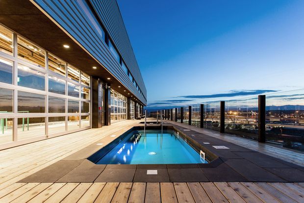 A rooftop terrace with a rectangular swimming pool beside a contemporary multi-storey building. The building features extensive floor-to-ceiling glazing, dark metal cladding, recessed exterior lighting, and a timber deck. A glass balustrade overlooks an urban landscape and distant mountains under a clear blue sky.