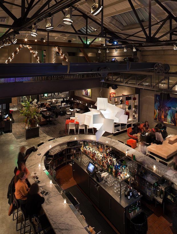 A dramatic interior view of a multi-level hospitality venue, likely a restaurant and bar, seen from an elevated mezzanine. The space features a large circular marble-topped bar with dark built-in cabinetry, dining and lounge seating, colourful artwork, sculptural white pendant lighting, exposed dark steel roof trusses, corrugated metal ceiling panels, and warm decorative string lights. The industrial structure contrasts with refined bar finishes and eclectic furnishings.