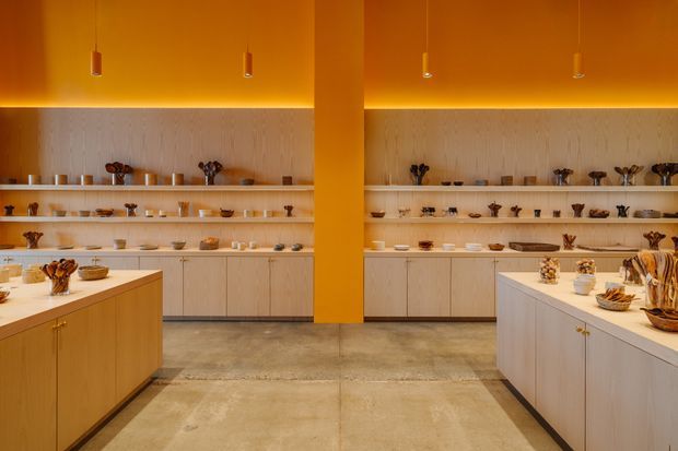 Warmly lit contemporary retail or display space with a central structural column, concrete flooring, pale timber built-in cabinetry, open shelving displaying ceramics and objects, and two large island counters in the foreground. Concealed linear lighting and small pendant lights create an amber, gallery-like atmosphere.