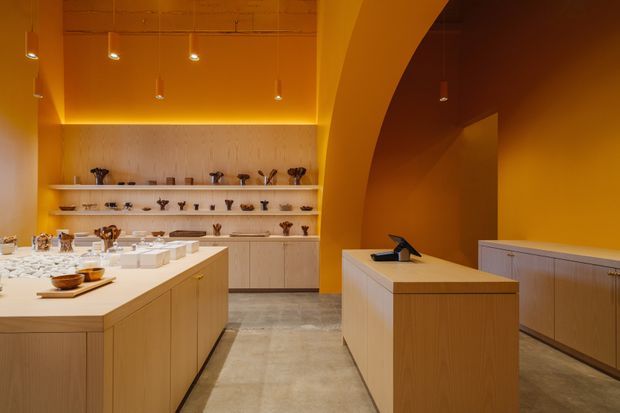 A warm, monochromatic retail or gallery-like interior with ochre-orange walls, ceiling, and a large curved archway. Light timber display cabinetry and central islands organize ceramics and small objects, while a textured pale feature wall with open shelving forms the rear display area. The space has polished concrete flooring and small suspended pendant lights, creating a refined, minimal exhibition atmosphere.