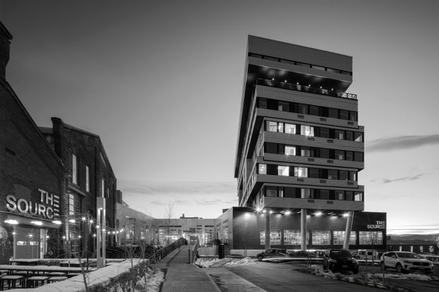 Black-and-white exterior architectural photograph of a contemporary multi-storey mixed-use building in an urban setting. The building has a strongly cantilevered and stepped upper form, horizontal bands, extensive glazing, rooftop or upper-level lighting, and a darker podium with commercial frontage. A landscaped pedestrian walkway, parking area, and adjacent older brick commercial buildings frame the scene. Visible signage reads “The Source.”