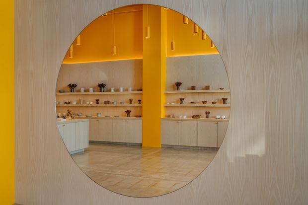 A warm, contemporary commercial interior viewed through a large circular opening in a pale timber wall. The space beyond features light timber cabinetry and open display shelving, a vivid mustard-yellow central structural wall and ceiling zone, small pendant lights, and a pale stone or concrete floor. The display area appears curated like a showroom, gallery, or retail space.