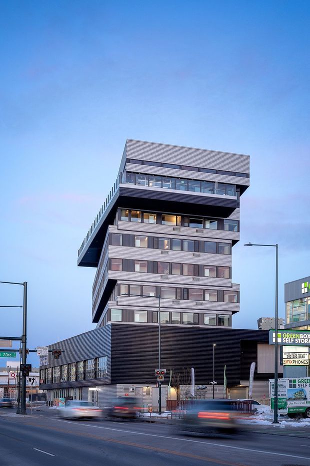 Exterior street-level photograph of a tall contemporary urban mixed-use building. The structure has stacked and projecting volumes, extensive horizontal metal cladding, dark lower-level cladding, broad glazed balconies, and numerous rectangular windows. It stands at a city intersection with streetlights, signage, traffic signals, and neighbouring commercial buildings visible around it.