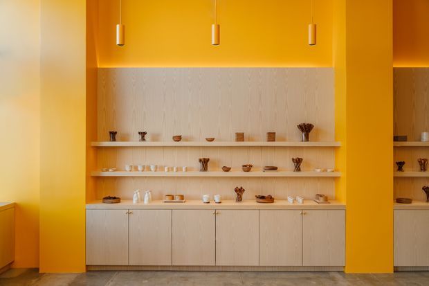 A warm, minimalist built-in display and storage wall with pale timber cabinetry, open shelving, and curated ceramics. The joinery is framed by vivid orange-yellow walls and tall matching panels, with slim pendant lights above and a grey stone or concrete-look floor below.