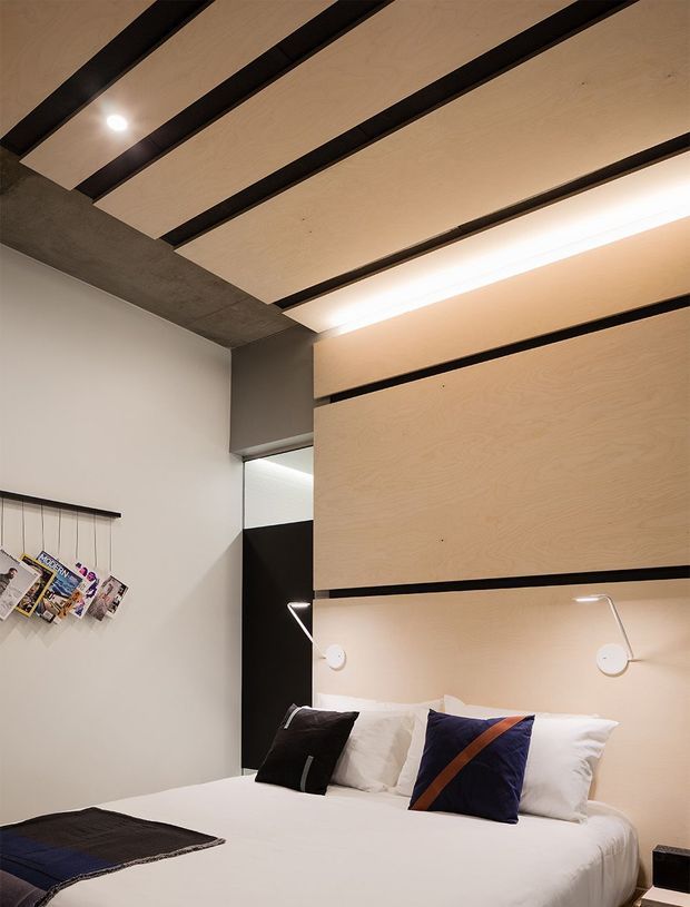 Compact contemporary bedroom with a white bed set against a built-in light timber feature wall and headboard. Black horizontal reveals and vertical panels create graphic contrast, while matching slim reading sconces flank the bed. The ceiling features suspended light timber panels with dark exposed gaps and integrated linear lighting. A small wall-mounted magazine display and a dark doorway or wardrobe opening are visible.