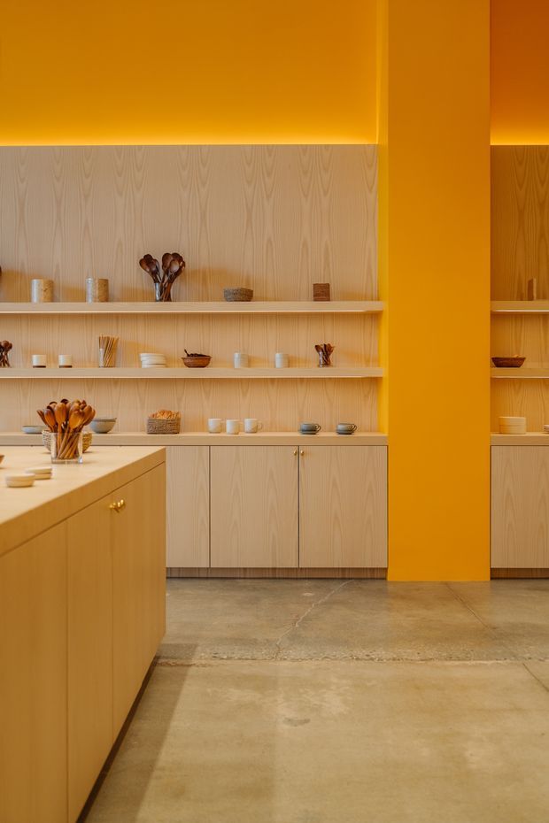Warm, contemporary kitchen interior with light timber cabinetry, open display shelving, a large island, and a vivid orange feature wall and column. The space has polished concrete flooring and integrated warm lighting, with minimalist ceramic and wooden objects arranged on the shelves.