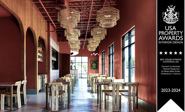 A long, narrow hospitality interior resembling a restaurant or bar, with deep burgundy-red walls and ceiling, exposed industrial roof structure, tall black-framed windows, arched timber-lined wall openings, and rows of light timber tables and chairs. Multiple sculptural tiered pendant lights hang centrally above the dining area, while a bar or service counter is visible toward the rear. A black awards panel occupies the right edge of the image.