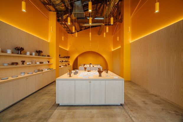 A spacious, warm-toned commercial showroom or retail interior with a central light-wood display island, open shelving filled with tableware, concrete flooring, vivid orange walls and ceiling planes, and numerous suspended pendant lights. The exposed ceiling services and structure create an industrial character, while the arched orange niche at the rear adds a sculptural focal point.